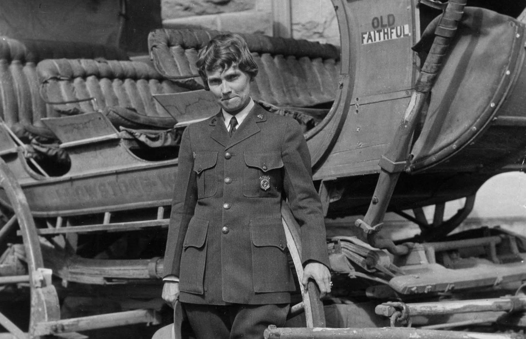 Herma Albertson poses in front of a stagecoach while wearing the NPS uniform with shield-shaped badge and holding a broad brim hat.